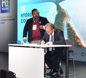 Two men at a hydrogen technology forum; one stands speaking, the other signs a document at a table.