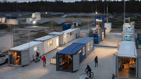 A view of several shipping containers converted to workspaces, with people walking and biking nearby at dusk. A view of several shipping containers converted to workspaces, with people walking and biking nearby at dusk.