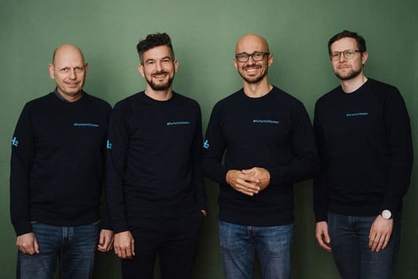 Four men in matching dark sweaters stand side by side, smiling, in front of a green background. Four men in matching dark sweaters stand side by side, smiling, in front of a green background.