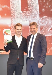Two men in suits smiling; one holds a trophy, standing in front of a red background with "H2" text.