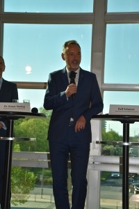 Man in a suit speaking into a microphone, standing by a table in front of large windows. Man in a suit speaking into a microphone, standing by a table in front of large windows.
