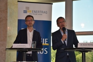 Two men in suits speak at a podium with microphones at an ENERTRAG event indoors. Two men in suits speak at a podium with microphones at an ENERTRAG event indoors.