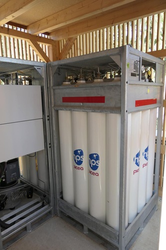 Rows of tall white gas cylinders labeled “HPS picea” in a metal storage unit under a wooden shelter. Rows of tall white gas cylinders labeled “HPS picea” in a metal storage unit under a wooden shelter.
