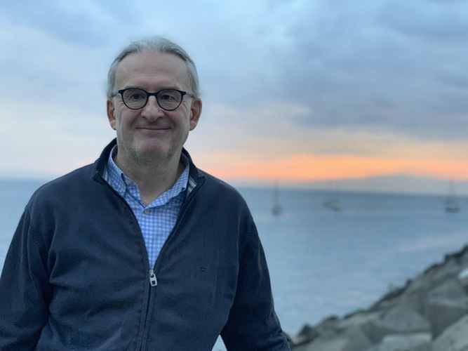 Smiling man in glasses stands by the sea at sunset, with sailboats and rocks in the background.