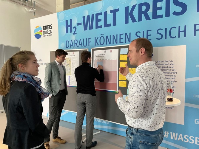 Four people discuss in front of an H2 World exhibition board with charts and notes in a modern indoor space. Four people discuss in front of an H2 World exhibition board with charts and notes in a modern indoor space.