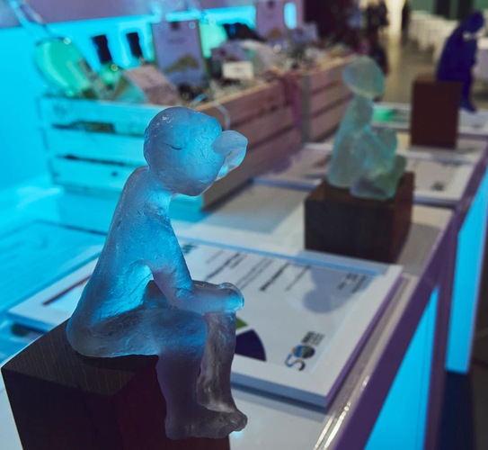 Small blue glass sculpture of a seated figure on a table with awards and gift baskets in the background. Small blue glass sculpture of a seated figure on a table with awards and gift baskets in the background.