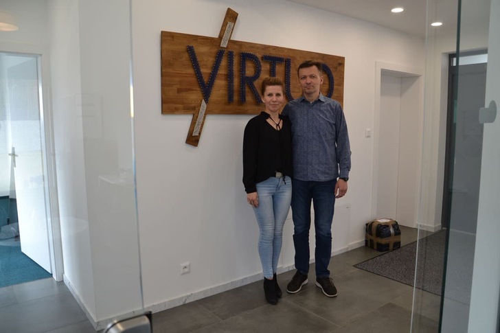 Two people stand side by side in an office hallway, with a "VIRTUS" sign on the wall behind them. Two people stand side by side in an office hallway, with a "VIRTUS" sign on the wall behind them.