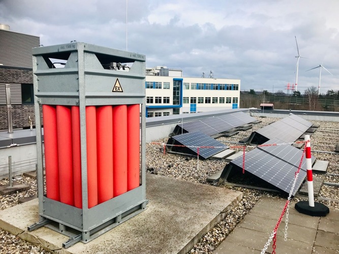 A rooftop with solar panels, a large red gas cylinder unit, and nearby office buildings in the background.