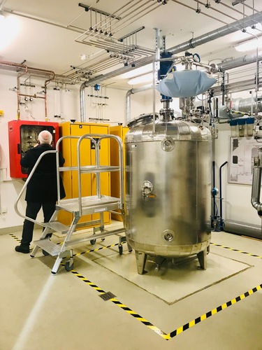 A person operates control panels in an industrial room with a large metal tank and safety markings on the floor.