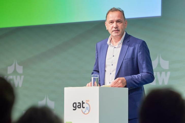 A man in a blue suit speaks at a podium labeled "gat 2023" with a glass of water on top. A man in a blue suit speaks at a podium labeled "gat 2023" with a glass of water on top.
