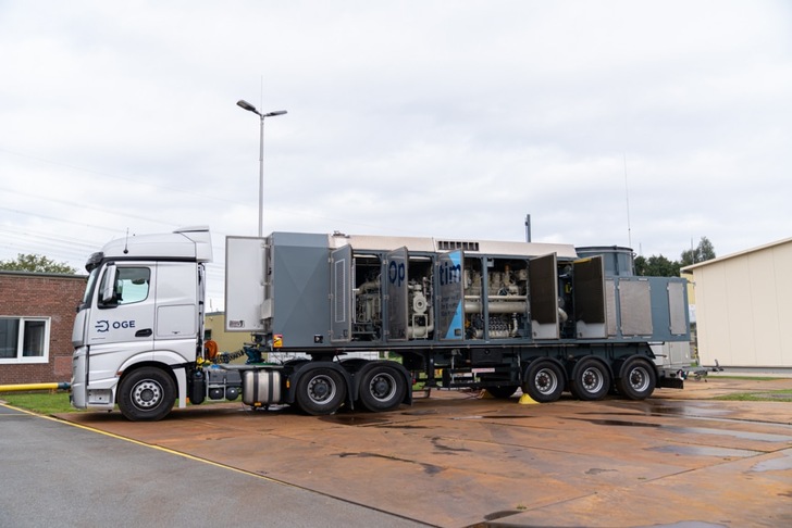 A large OGE truck with an open-sided trailer showing industrial equipment parked outside a building. A large OGE truck with an open-sided trailer showing industrial equipment parked outside a building.