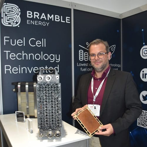 A man in a suit holds a fuel cell at a Bramble Energy booth displaying fuel cell technology. A man in a suit holds a fuel cell at a Bramble Energy booth displaying fuel cell technology.