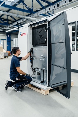 A technician kneels, inspecting an open industrial machine labeled "picea" in a factory setting. A technician kneels, inspecting an open industrial machine labeled "picea" in a factory setting.