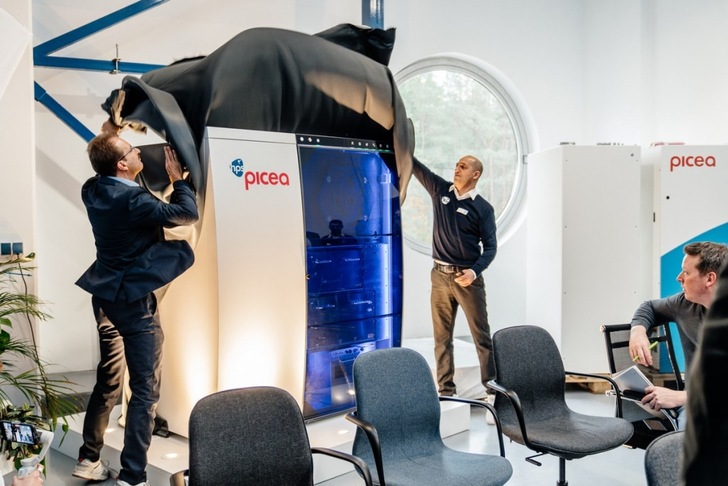 Two men unveil a machine labeled "picea" in front of an audience in a modern, bright room. Two men unveil a machine labeled "picea" in front of an audience in a modern, bright room.