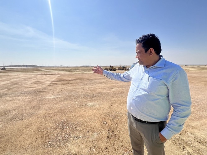 A man in a button-up shirt gestures toward an open, barren landscape under a clear sky. A man in a button-up shirt gestures toward an open, barren landscape under a clear sky.
