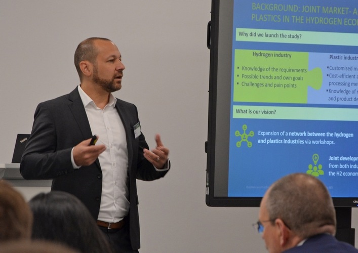 A man in a suit presents slides about hydrogen and plastics to an audience in a conference room.
