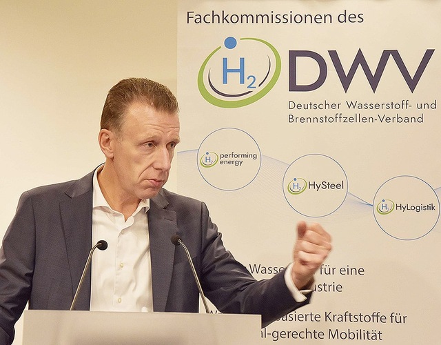 A man in a suit gestures while speaking at a podium with a DWV hydrogen energy banner behind him. A man in a suit gestures while speaking at a podium with a DWV hydrogen energy banner behind him.