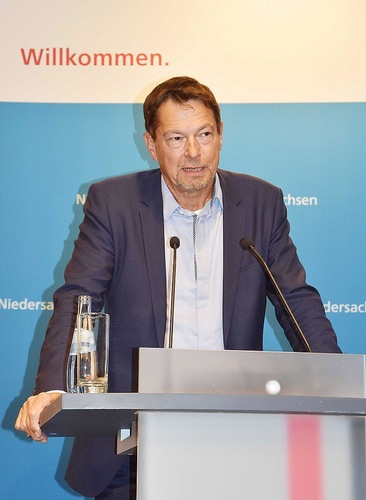 A man in a suit speaks at a podium with microphones; "Willkommen" is written on the wall behind him. A man in a suit speaks at a podium with microphones; "Willkommen" is written on the wall behind him.