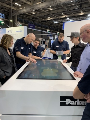 Six people stand around a large touchscreen table at a trade show, engaging in a technology demonstration. Six people stand around a large touchscreen table at a trade show, engaging in a technology demonstration.