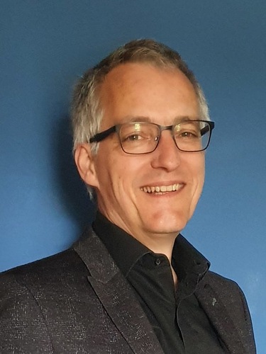 A smiling man with glasses and gray hair wearing a dark jacket, standing in front of a blue background. A smiling man with glasses and gray hair wearing a dark jacket, standing in front of a blue background.
