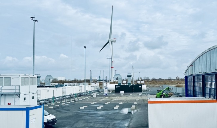 A wind turbine stands behind white industrial buildings and equipment under a cloudy sky. A wind turbine stands behind white industrial buildings and equipment under a cloudy sky.