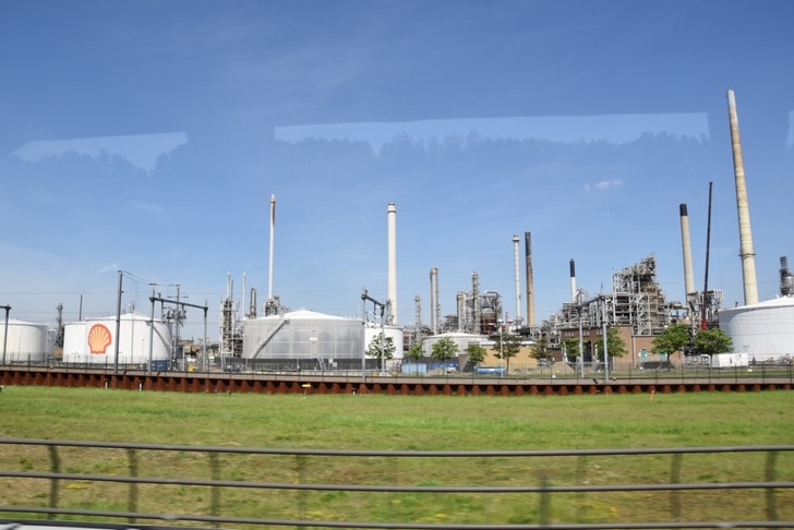 Oil refinery with storage tanks, chimneys, and a Shell logo under a clear blue sky, viewed from a distance.