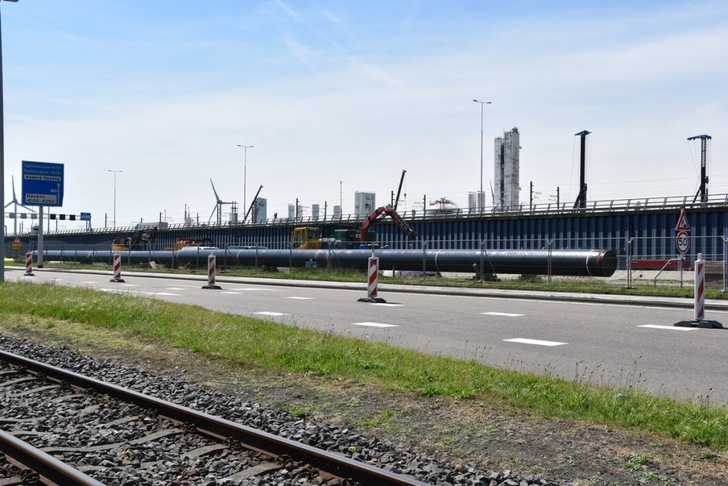 Large industrial pipes and construction barriers line a road near a railway track and industrial buildings. Large industrial pipes and construction barriers line a road near a railway track and industrial buildings.