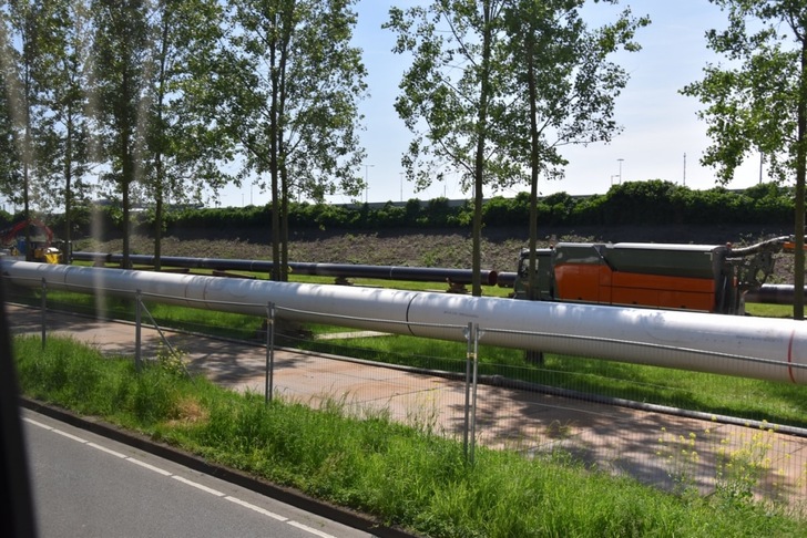 Large metal pipeline runs beside a road and trees under a clear sky, with machinery in the background.