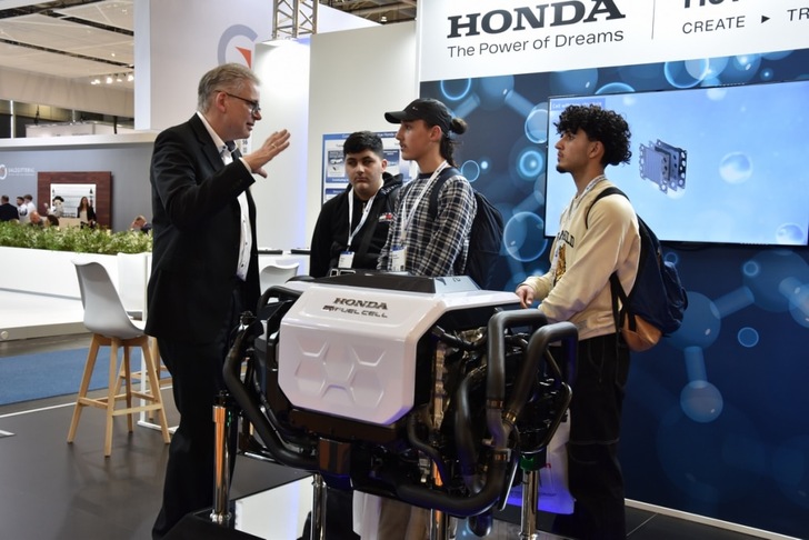 A man explains a Honda fuel cell engine to three young men at a technology exhibition booth. A man explains a Honda fuel cell engine to three young men at a technology exhibition booth.