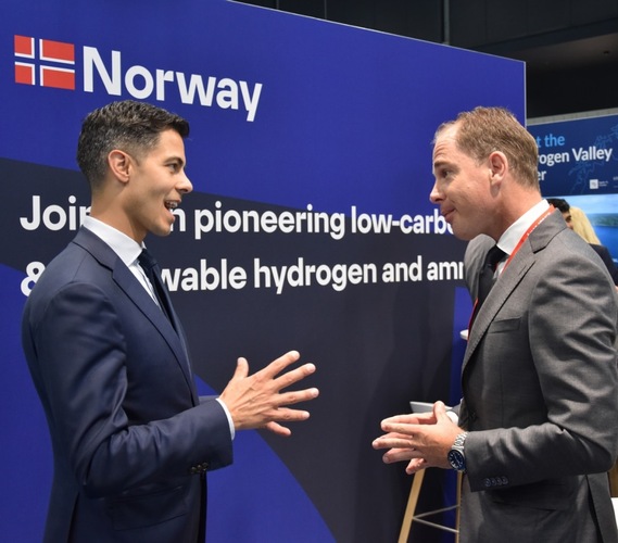 Two men in suits have a conversation in front of a Norway booth about hydrogen and ammonia solutions. Two men in suits have a conversation in front of a Norway booth about hydrogen and ammonia solutions.