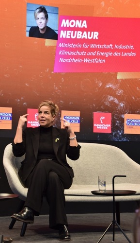 Mona Neubaur speaks on stage at a conference, seated in front of a screen displaying her name and title. Mona Neubaur speaks on stage at a conference, seated in front of a screen displaying her name and title.