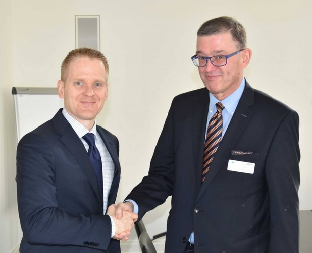 Two men in suits shaking hands and smiling in an office setting.