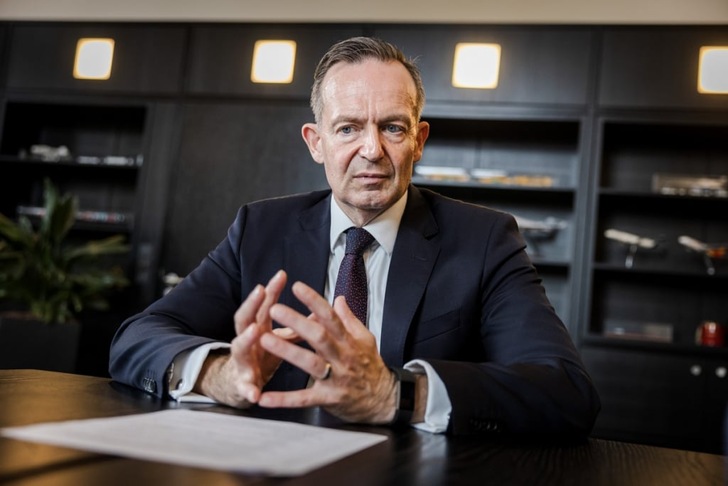 Man in a suit sitting at a desk with papers, hands clasped, bookshelves in the background.