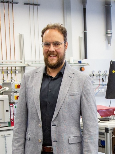 A man with glasses and a beard, wearing a gray blazer, stands in a laboratory setting. A man with glasses and a beard, wearing a gray blazer, stands in a laboratory setting.