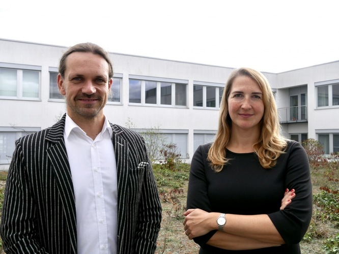 Two people standing outside a modern white building, both looking at the camera and smiling.