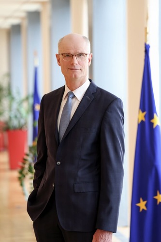 Man in a suit and tie stands indoors next to an EU flag, with plants and windows in the background. Man in a suit and tie stands indoors next to an EU flag, with plants and windows in the background.