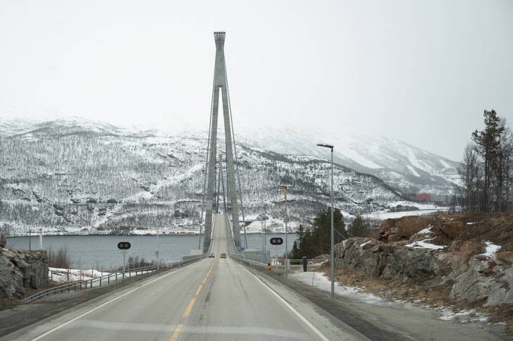 A suspension bridge crosses a snowy river valley with mountains in the background, viewed from the road.
