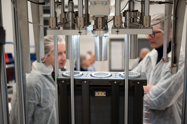 Two people in lab coats work near industrial equipment with metal cylinders in a laboratory setting. Two people in lab coats work near industrial equipment with metal cylinders in a laboratory setting.