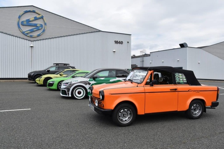 Four cars, including an orange convertible, are parked in a row outside a modern industrial building.