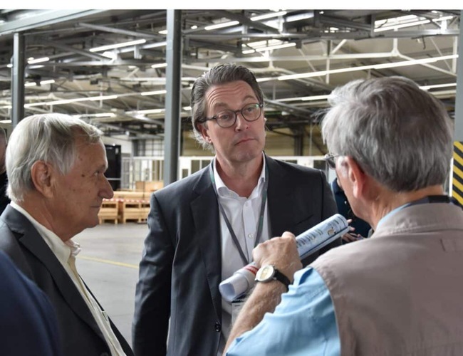 Three men in conversation inside an industrial building, one holding rolled-up papers, with shelves in the background.