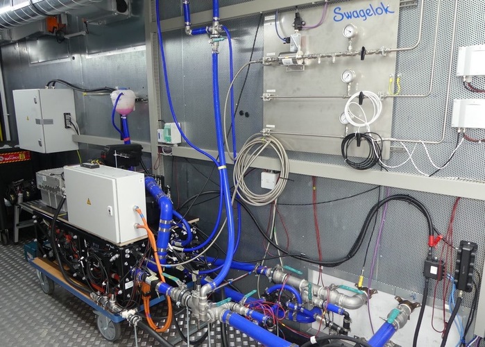 Industrial lab setup with blue pipes, control panels, gauges, and labeled Swagelok wall-mounted equipment. Industrial lab setup with blue pipes, control panels, gauges, and labeled Swagelok wall-mounted equipment.