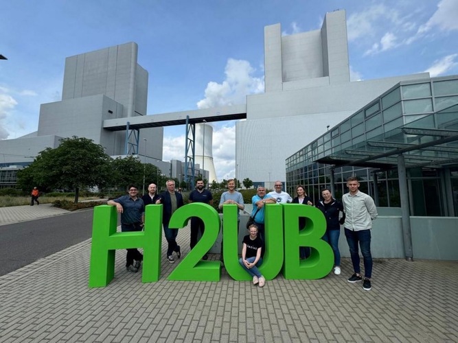 A group of people poses outdoors with large green "H2UB" letters in front of industrial buildings. A group of people poses outdoors with large green "H2UB" letters in front of industrial buildings.