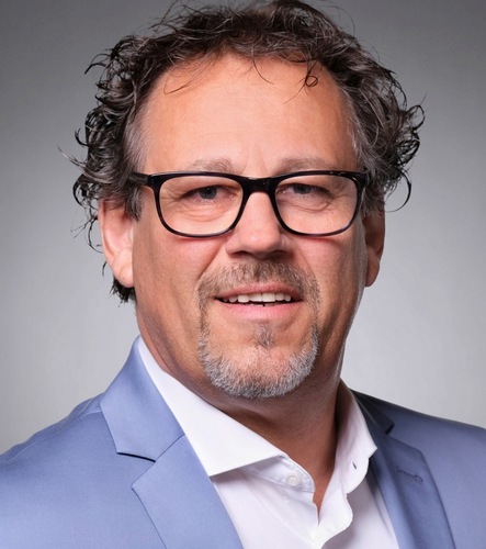 Smiling middle-aged man with glasses, curly hair, and a blue suit jacket over a white shirt.