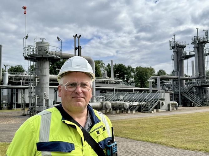 Man in safety gear and hard hat stands in front of industrial facility with pipes and tanks outdoors.