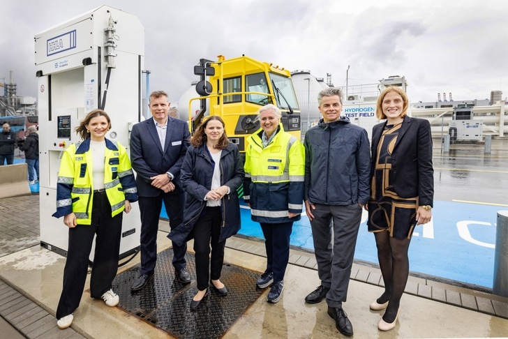 Six people in business attire and safety jackets stand by a hydrogen fueling station and truck on a wet day. Six people in business attire and safety jackets stand by a hydrogen fueling station and truck on a wet day.