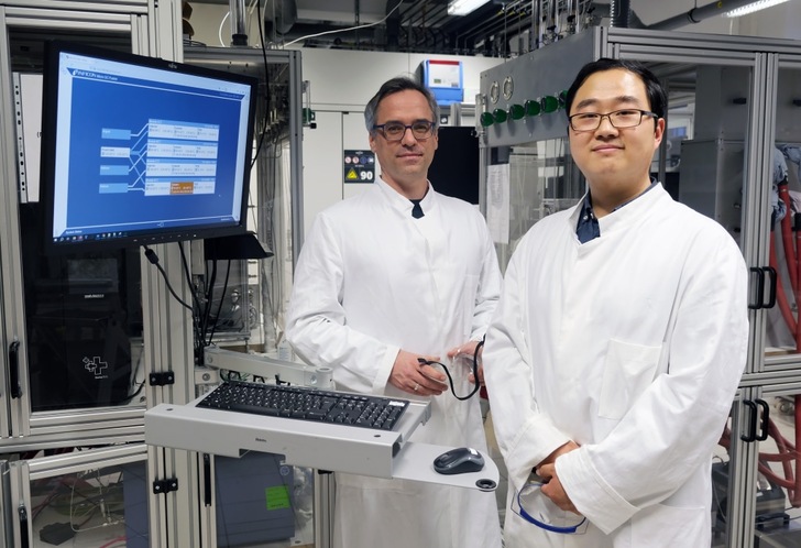 Two scientists in white lab coats stand by lab equipment and a computer displaying data. Two scientists in white lab coats stand by lab equipment and a computer displaying data.