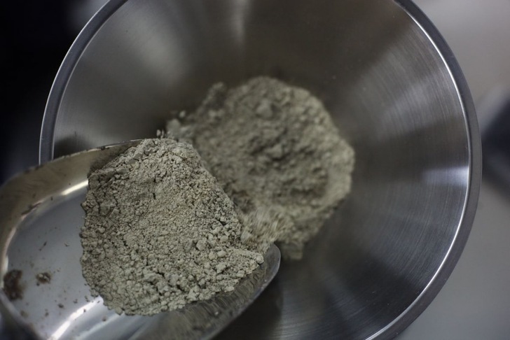 A metal scoop pours gray powder into a stainless steel mixing bowl. A metal scoop pours gray powder into a stainless steel mixing bowl.