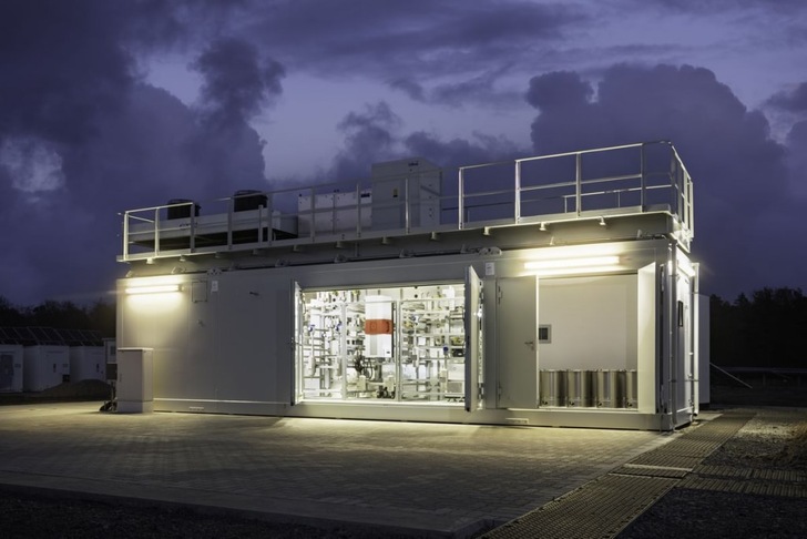 Industrial facility lit up at dusk, with visible machinery inside and dramatic clouds in the background.