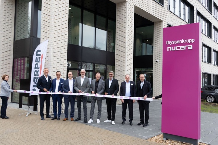 Eight people stand outside a building cutting a ribbon near a sign reading "thyssenkrupp nucera. Eight people stand outside a building cutting a ribbon near a sign reading "thyssenkrupp nucera.