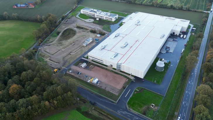 Aerial view of a large industrial building with surrounding roads, fields, and nearby trees. Aerial view of a large industrial building with surrounding roads, fields, and nearby trees.
